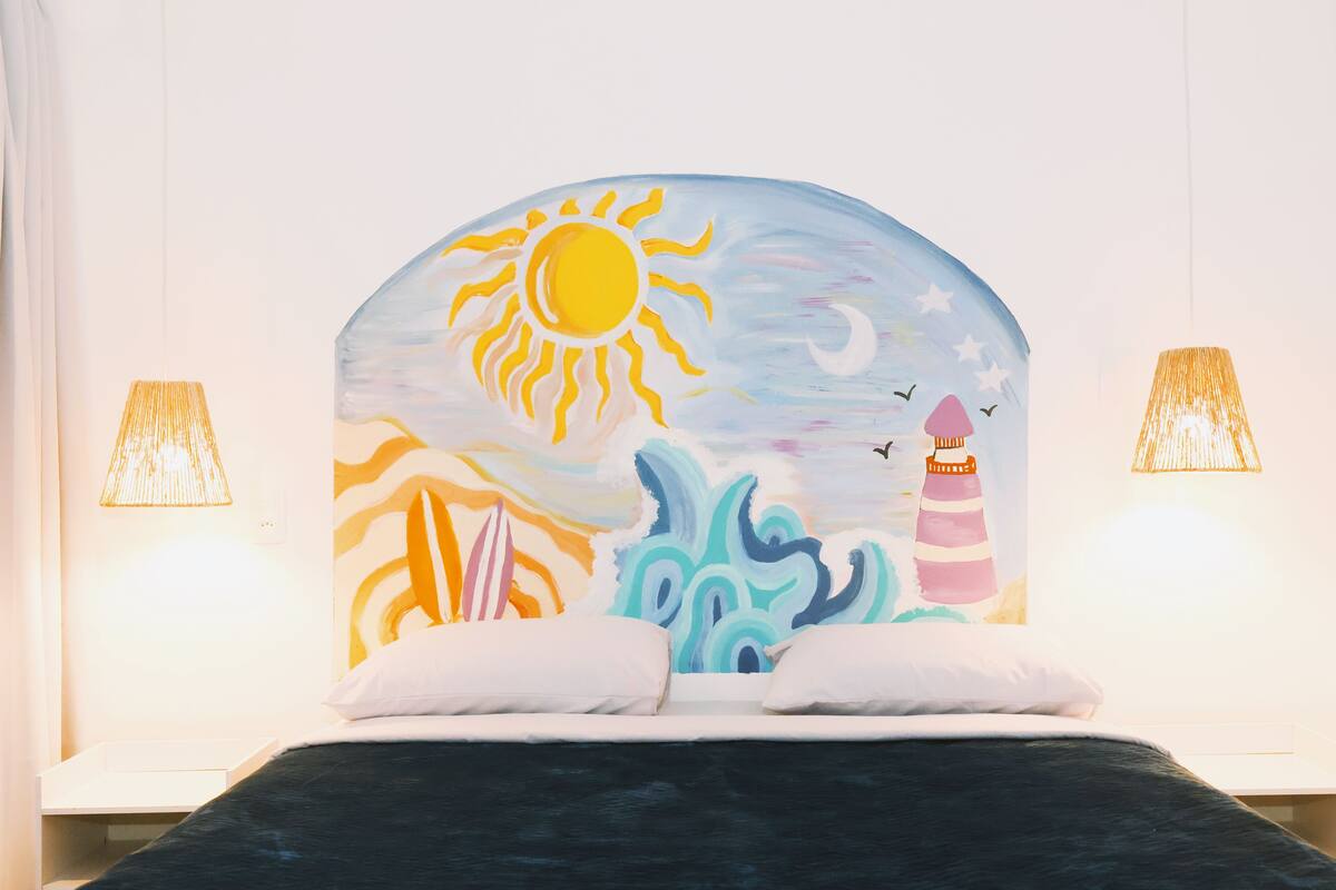 Cama com um mural pintado representando um sol, ondas e um farol. Há duas luminárias de cada lado da cama.
