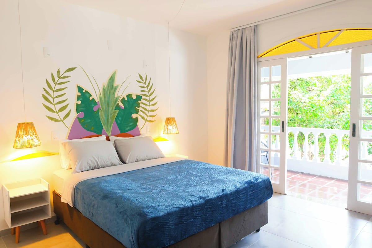 Quarto com cama, luminárias de cabeceira, uma janela grande e vista para o verde lá fora.