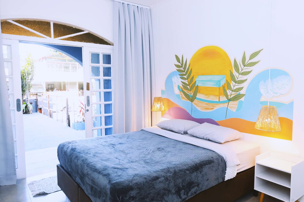 Quarto com cama, mesa de cabeceira, painel decorativo na parede e vista para o exterior.