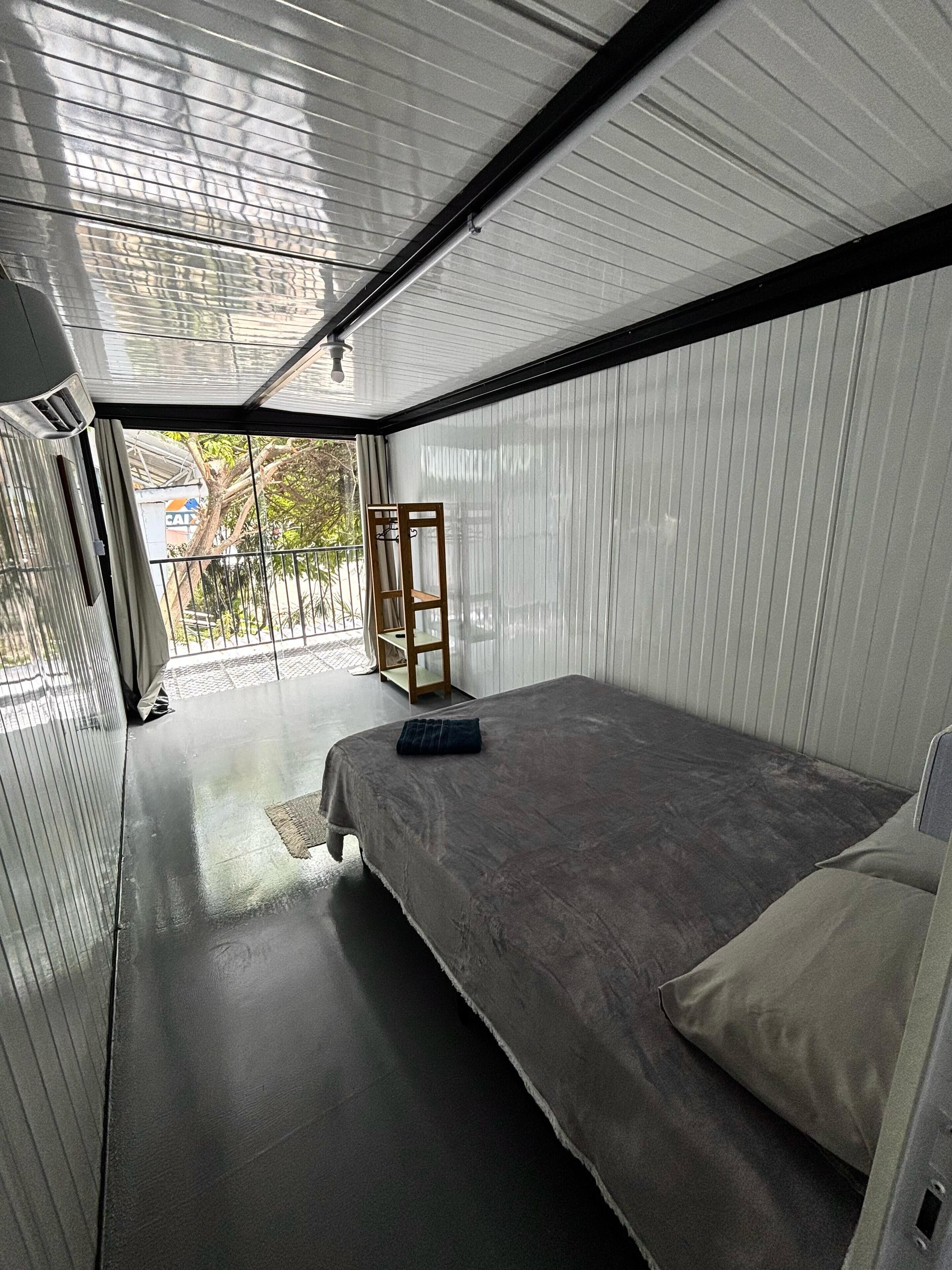 A compact, modern room with a bed, a small table, and a chair. The room has a slanted ceiling and a view of the outside.