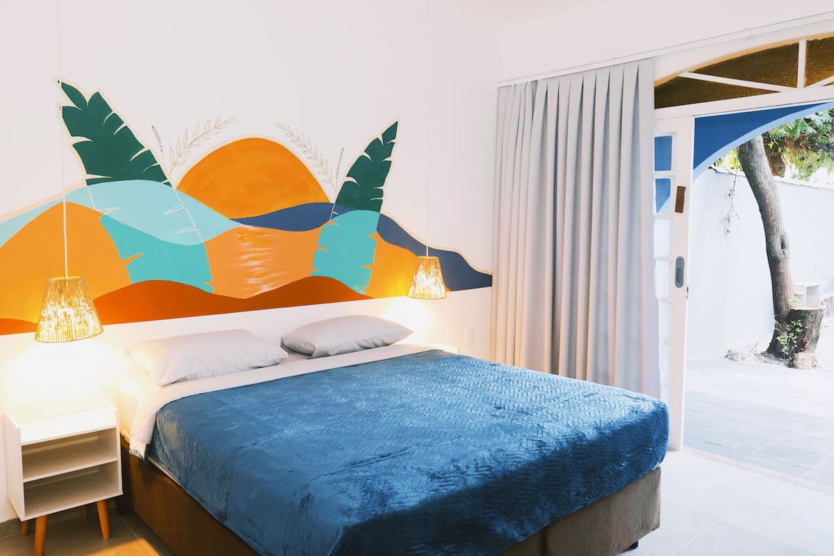 Quarto com cama, mesa de cabeceira, painel decorativo na parede e vista para o exterior.