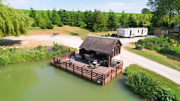 Een houten terras met buiten zitplaatsen en een klein gebouw, omgeven door groen en water.