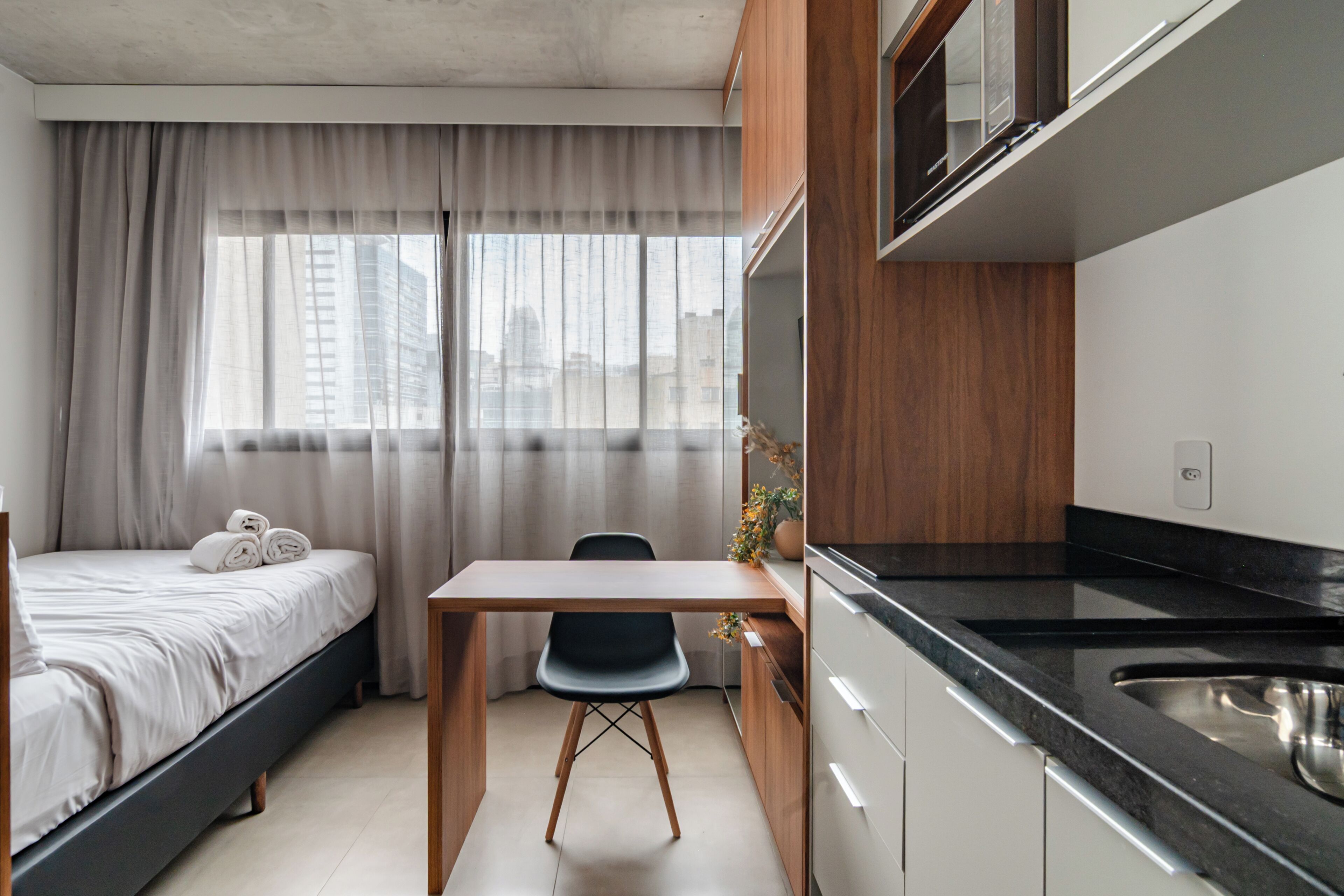 Een kleine hotelkamer met een bed, bureau, stoel, kitchenette en raam met gordijnen.