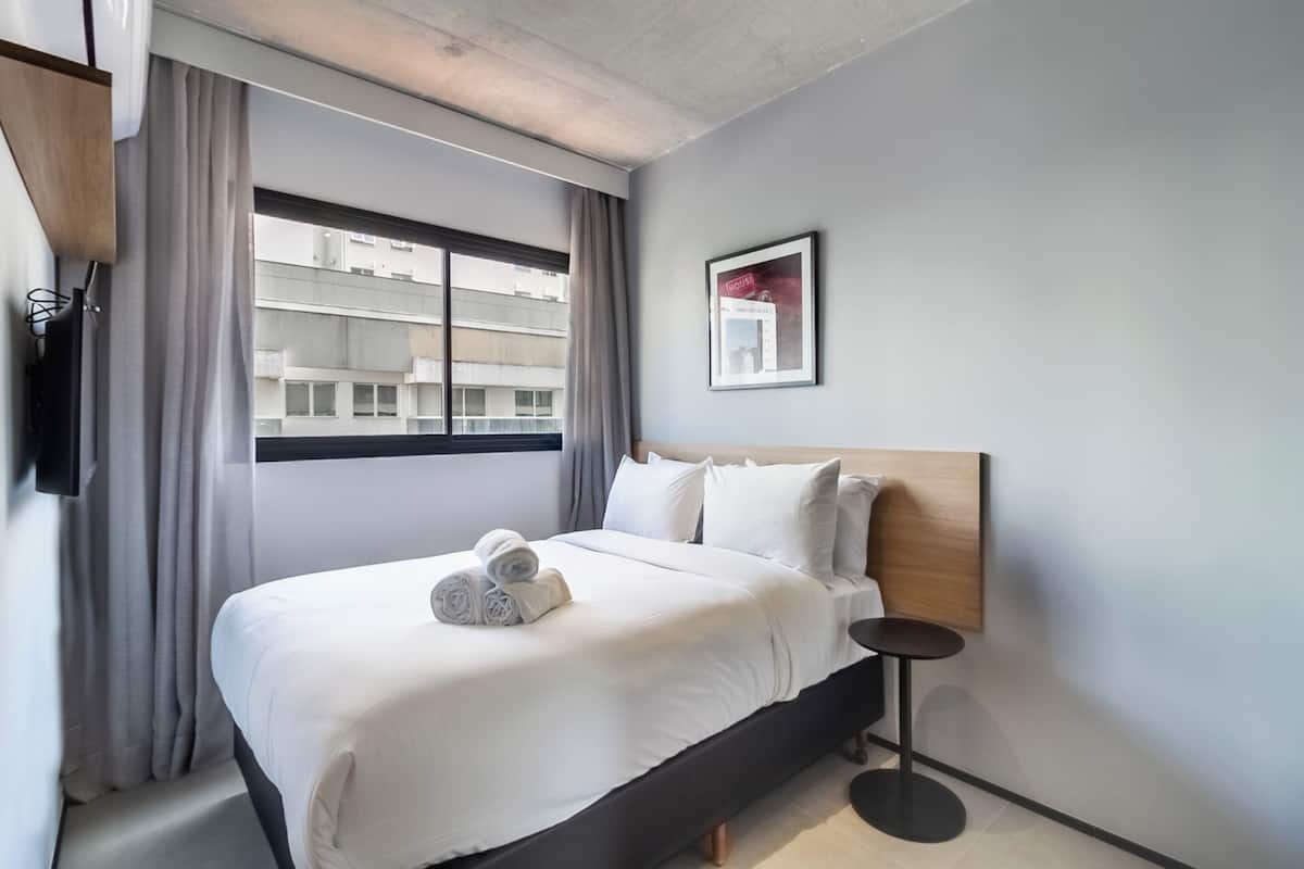 Une chambre d’hôtel moderne avec un grand lit, une petite table de chevet, une fenêtre avec des rideaux et un tableau encadré au mur.