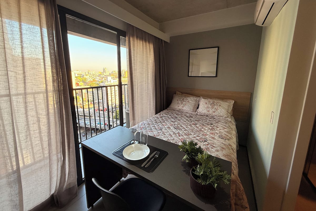 Une chambre d’hôtel avec un lit, un bureau et une chaise, une petite plante en pot et une fenêtre avec des rideaux.