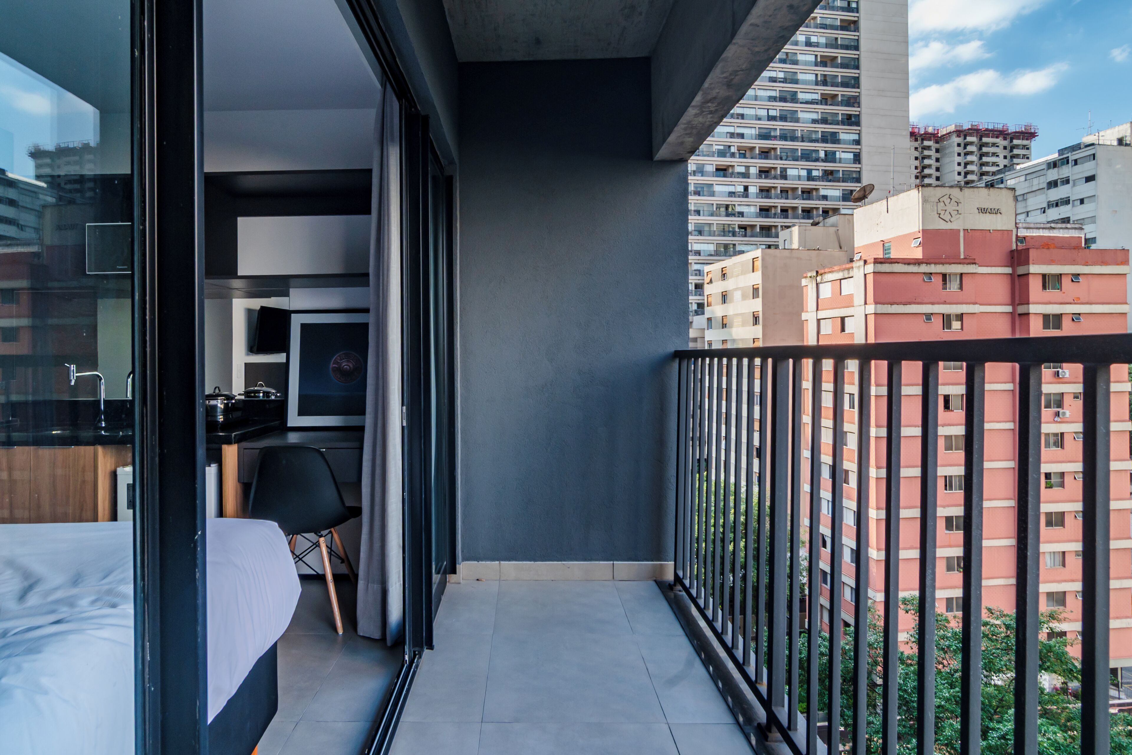 Uma varanda com cama, uma cadeira, área de cozinha e vista para edifícios ao redor.