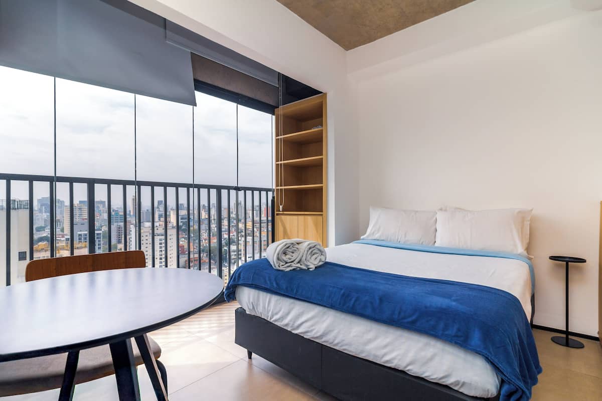 Une chambre moderne avec une grande fenêtre, un lit recouvert d’une couette bleue et blanche, une table de chevet en bois et une petite table ronde.