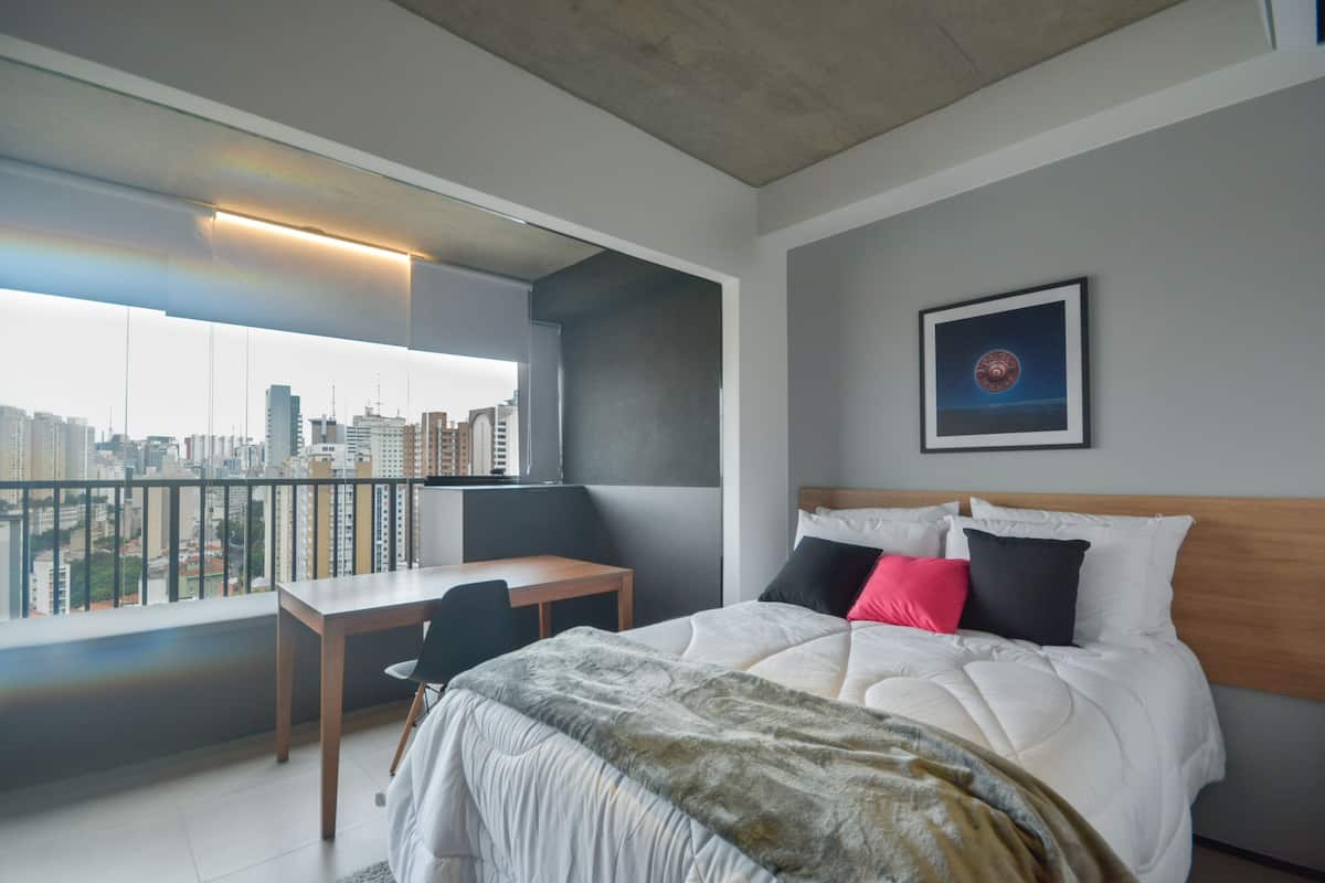 Une chambre d’hôtel moderne avec vue sur la ville, un balcon, un lit avec plusieurs oreillers et un petit bureau.