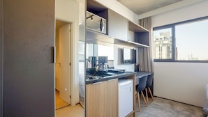 Cocina compacta con refrigerador integrado, cafetera y un lavamanos pequeño. Hay una zona de comedor con mesa y sillas, y un ventanal con vistas a la ciudad.
