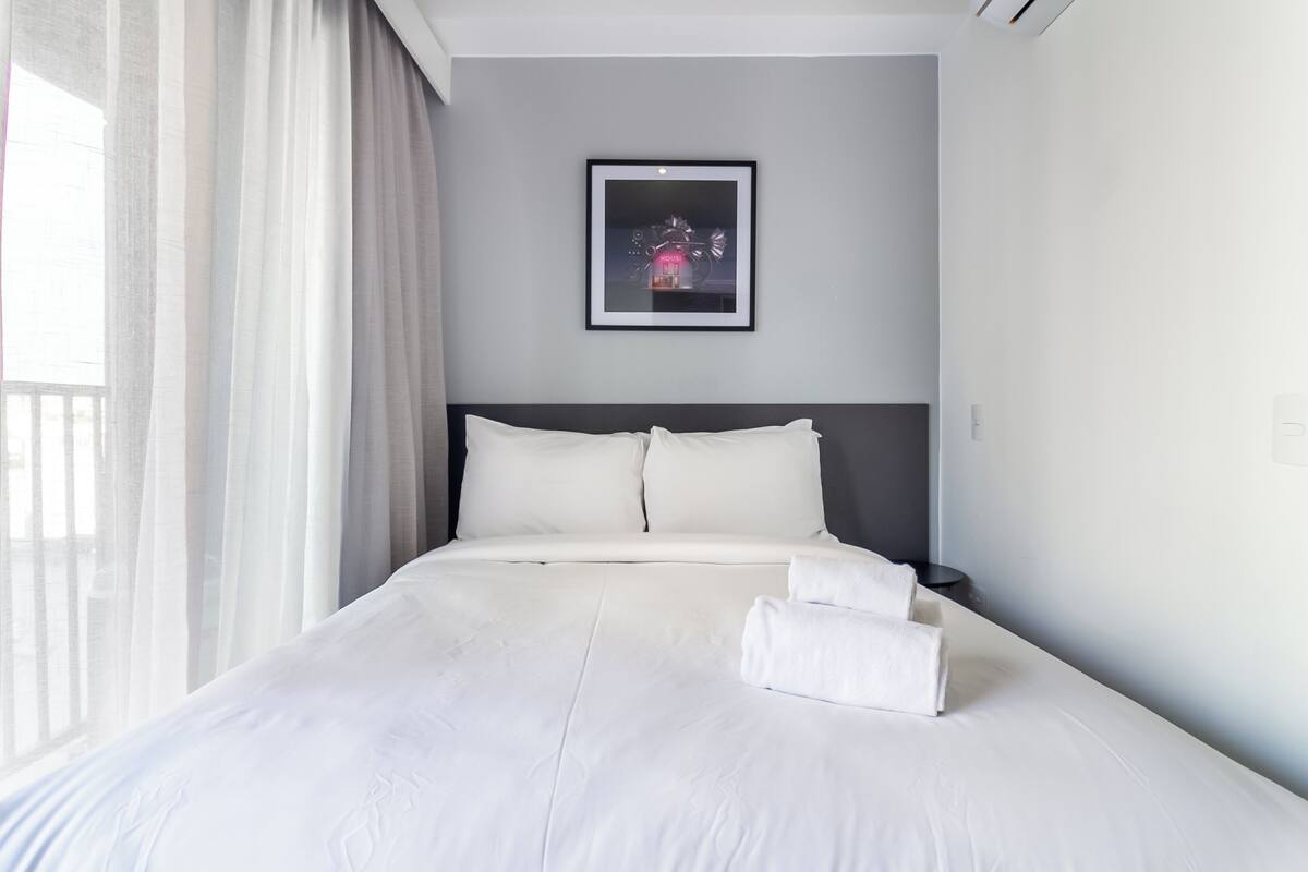 Un lit bien fait, avec du linge de lit blanc et des oreillers, un tableau encadré au mur et une fenêtre ornée de rideaux légers.