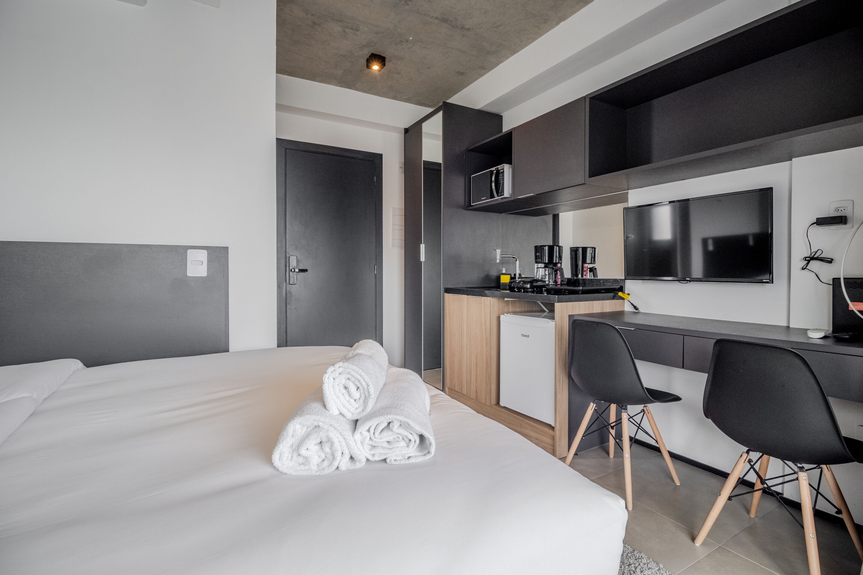 Habitación de hotel moderna con una cama bien hecha, una kitchenette pequeña, un escritorio con dos sillas y un televisor montado sobre la encimera.