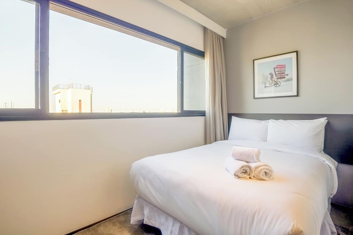 Une chambre d’hôtel avec une grande fenêtre, un lit recouvert de draps blancs et un tableau encadré au mur.