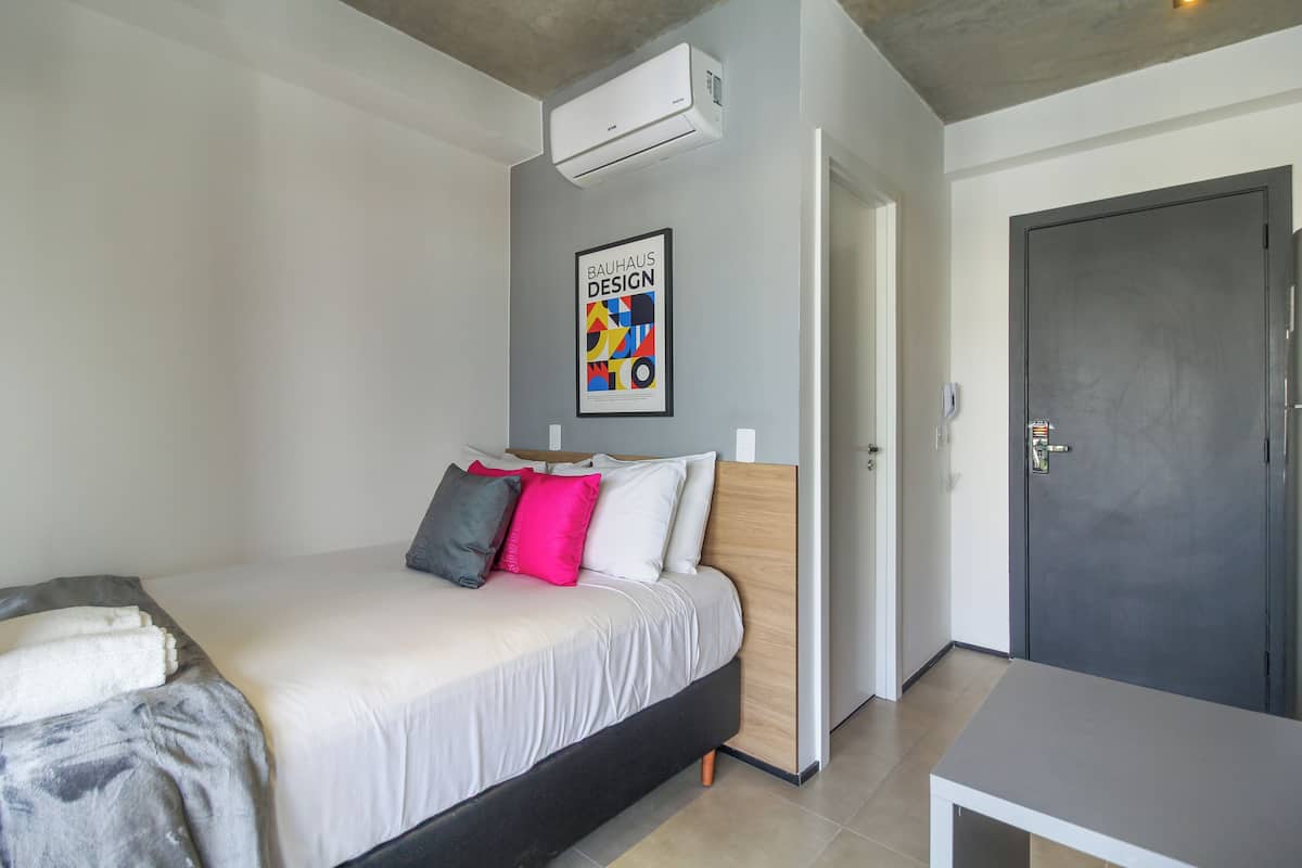 Une chambre à coucher moderne avec un lit, des oreillers, une affiche encadrée, un climatiseur et une porte.