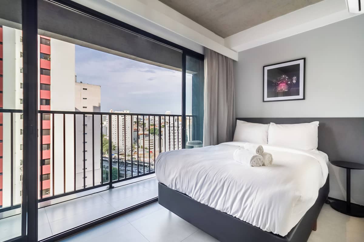 Une chambre d’hôtel moderne avec un grand lit, une vue sur la ville depuis un balcon et un tableau encadré au mur.