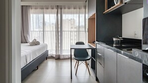 Habitación de hotel pequeña con cama, una mesa de comedor con silla, zona de cocina con microondas y vistas por la ventana.
