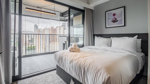 Un cuarto de hotel con una cama grande, vista de la ciudad a través de un ventanal corredero y un cuadro en la pared.
