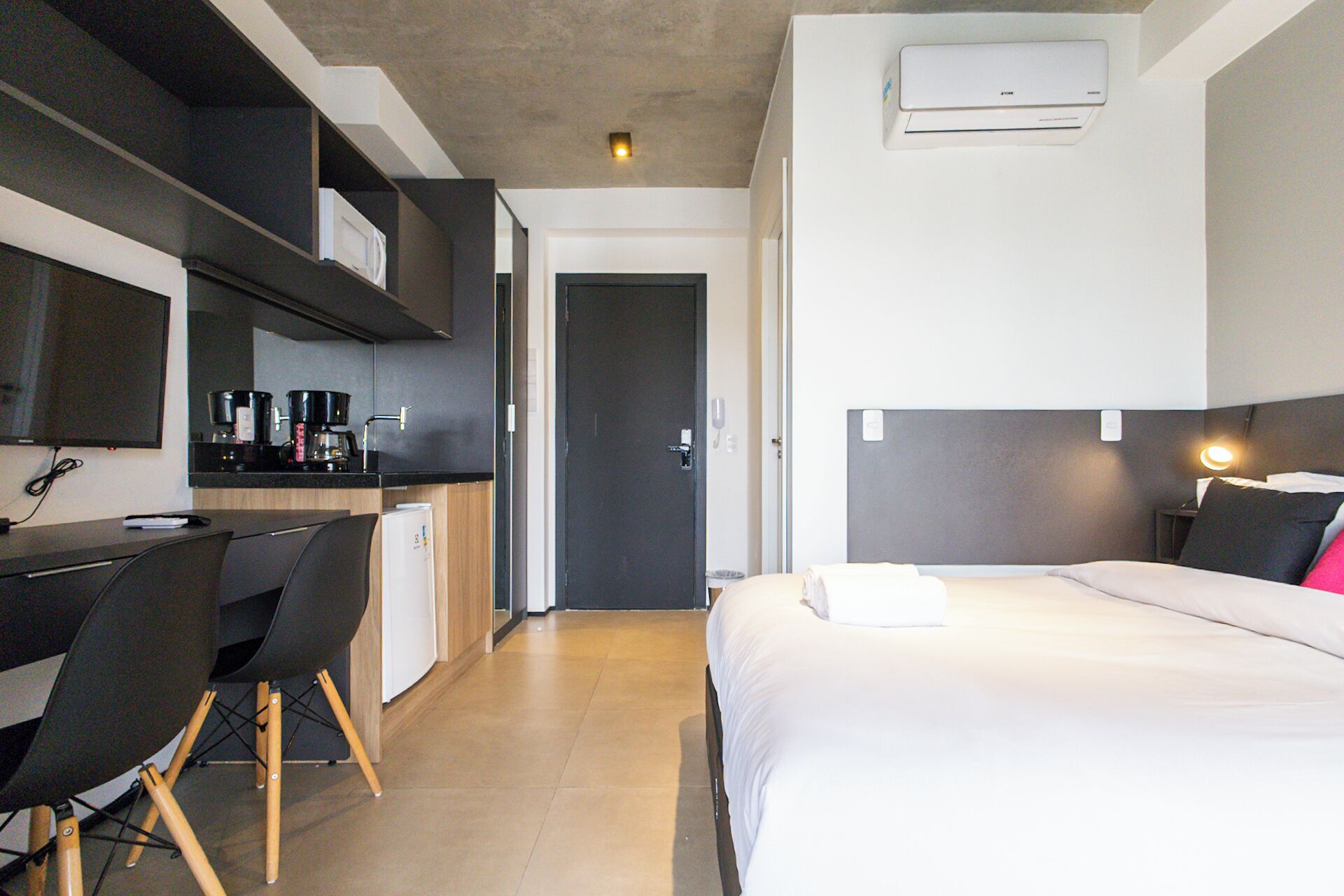 A modern hotel room with a bed, a desk, and a kitchenette.