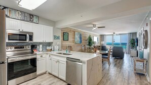 A modern kitchen with white cabinets, a built-in oven, and a microwave. There is a dining area with a table and chairs, and a view of the ocean through large windows. - Pelican Beach 1213 2 Bedroom Condo (Destin)