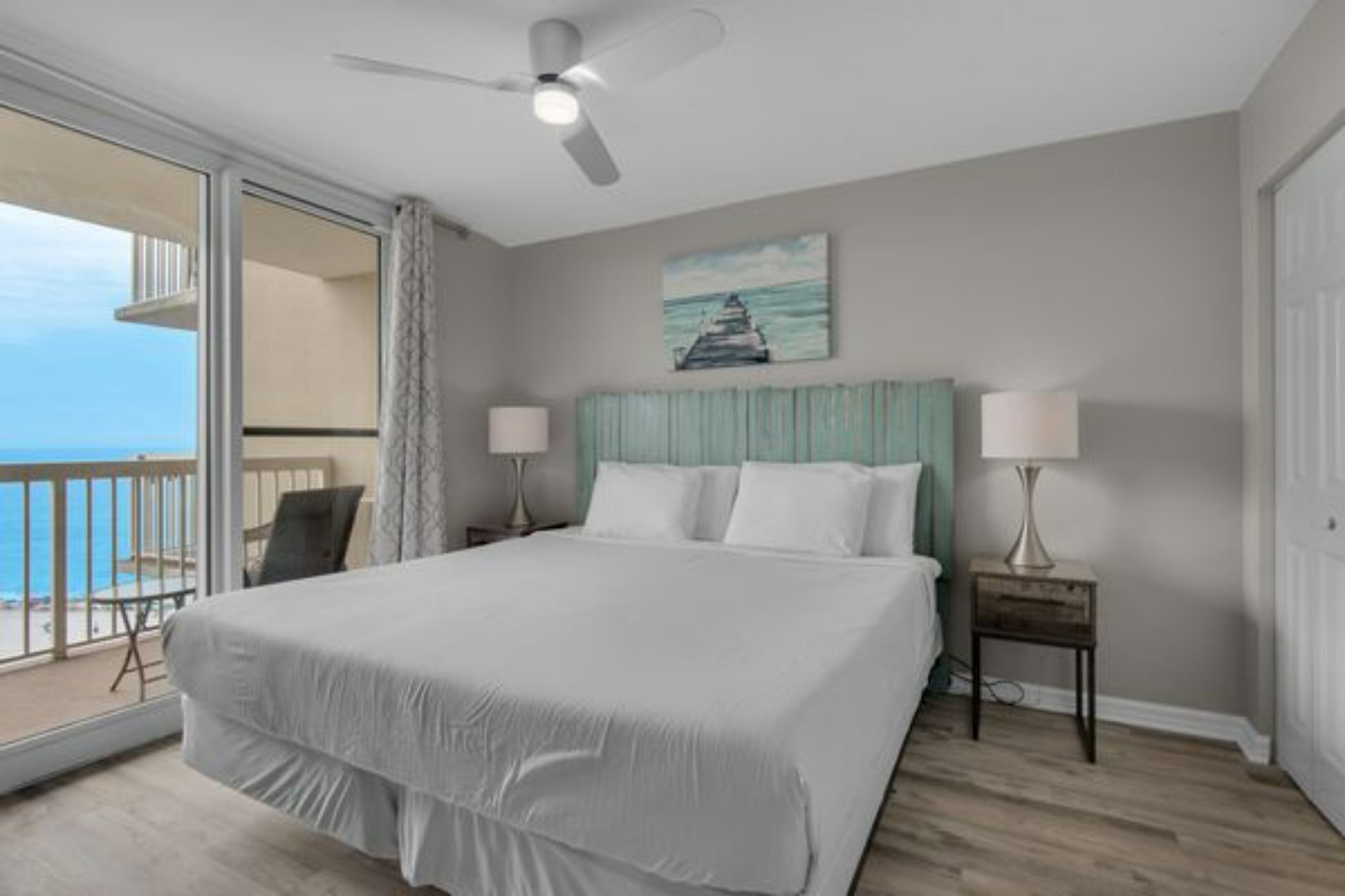 A hotel room with a bed, a ceiling fan, a painting of a lighthouse, and a view of the ocean through a balcony.