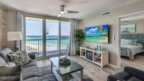 A modern living room with a large TV, a balcony with a view of the beach, and a cozy seating area. - Pelican Beach 1213 2 Bedroom Condo (Destin)