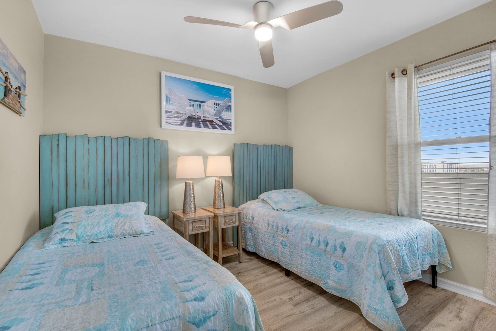 A bedroom with two beds, a ceiling fan, a nightstand with lamps, a painting, and a window with blinds.