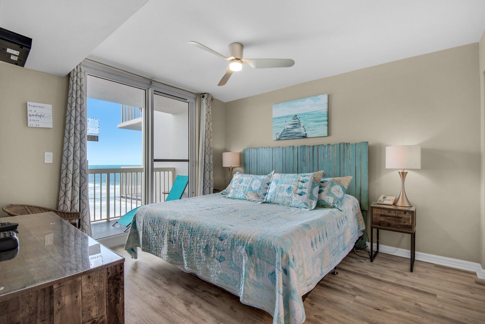 A bedroom with a bed, a desk, a chair, a nightstand, and a view of the ocean.