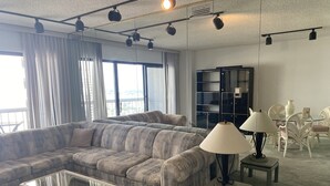 Ein modernes Wohnzimmer mit einem Ecksofa, einem Glastisch und einem kleinen Beistelltisch mit Lampe. Es gibt einen Esstisch mit Glasplatte und Stühlen sowie eine schwarze Kommode an der Wand.
