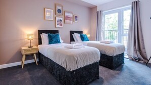 A hotel room with two beds, a small bedside table with a lamp, framed pictures on the wall, and a window with curtains.