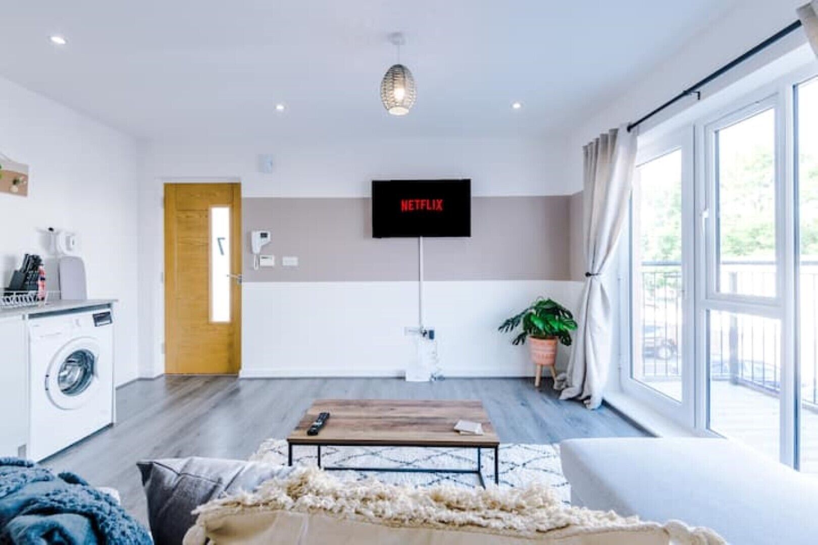A modern living room with a washing machine, a mounted TV displaying 'NETFLIX', a wooden coffee table, and a large window with curtains.