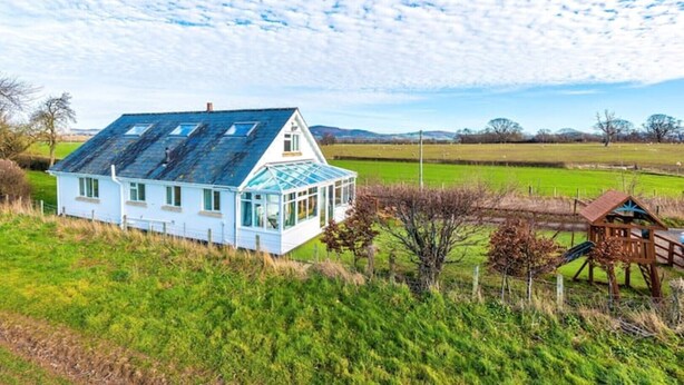 Une maison blanche dotée d’une véranda en verre, entourée de champs verdoyants et agrémentée d’une structure de jeux en bois.