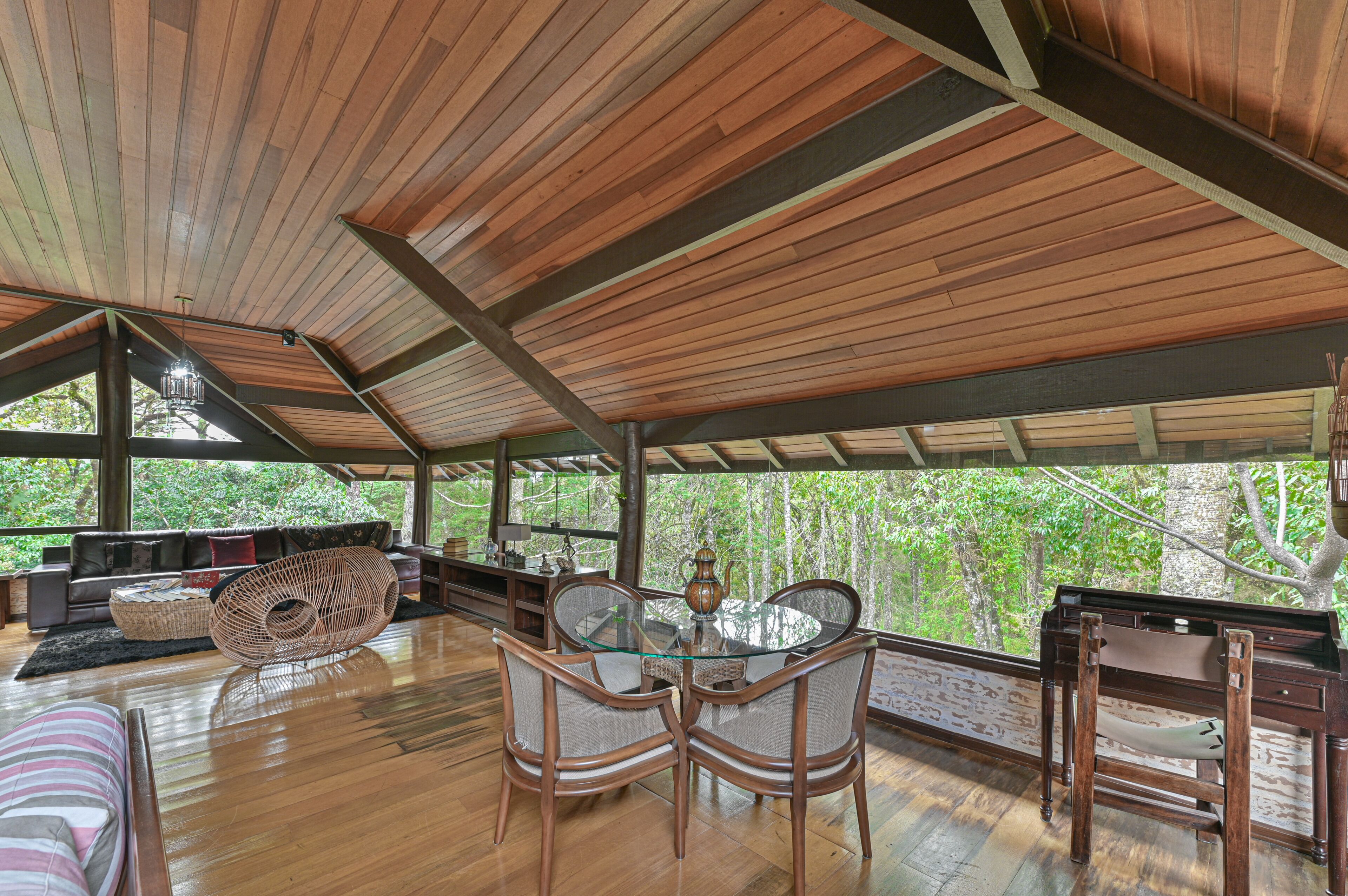 A spacious room with a wooden ceiling, a glass table with chairs, a sofa, and a view of trees through large windows.