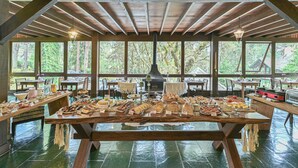 A large wooden table with various food items and a wood stove in a covered dining area with large windows.