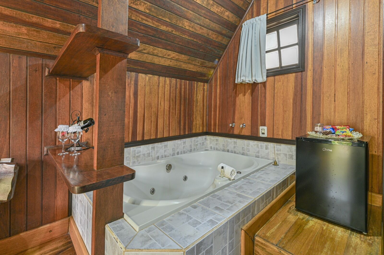 A wooden cabin bathroom with a large bathtub, a small refrigerator, and a wooden shelf with glasses and a bottle.