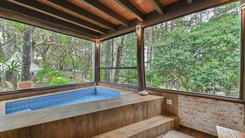 Zona de spa con un banco de madera, una pequeña piscina y vistas a un área boscosa a través de grandes ventanales.