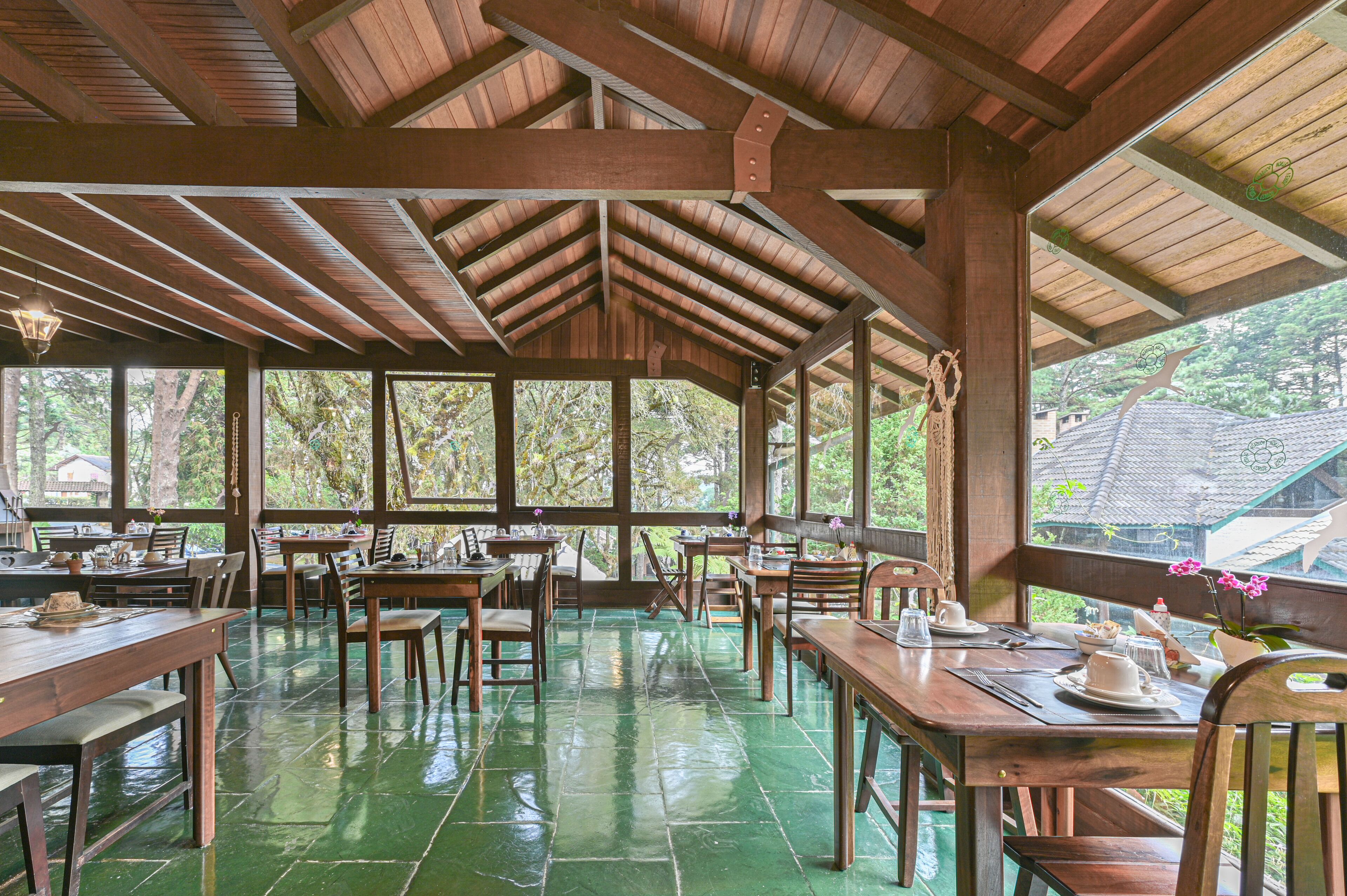 Un área de comedor cubierta con mesas y sillas de madera, ventanas amplias y un suelo de baldosas verdes.