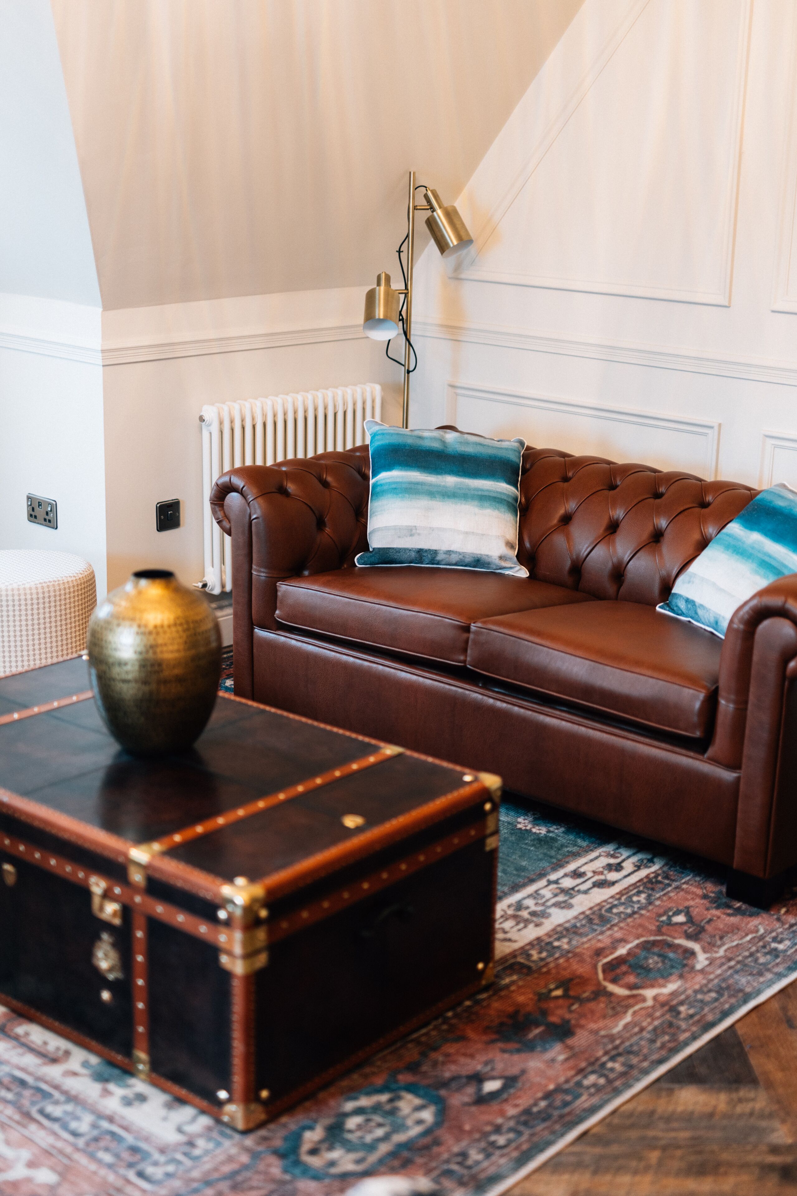 A brown leather sofa with striped pillows, a vintage trunk, and a floor lamp.