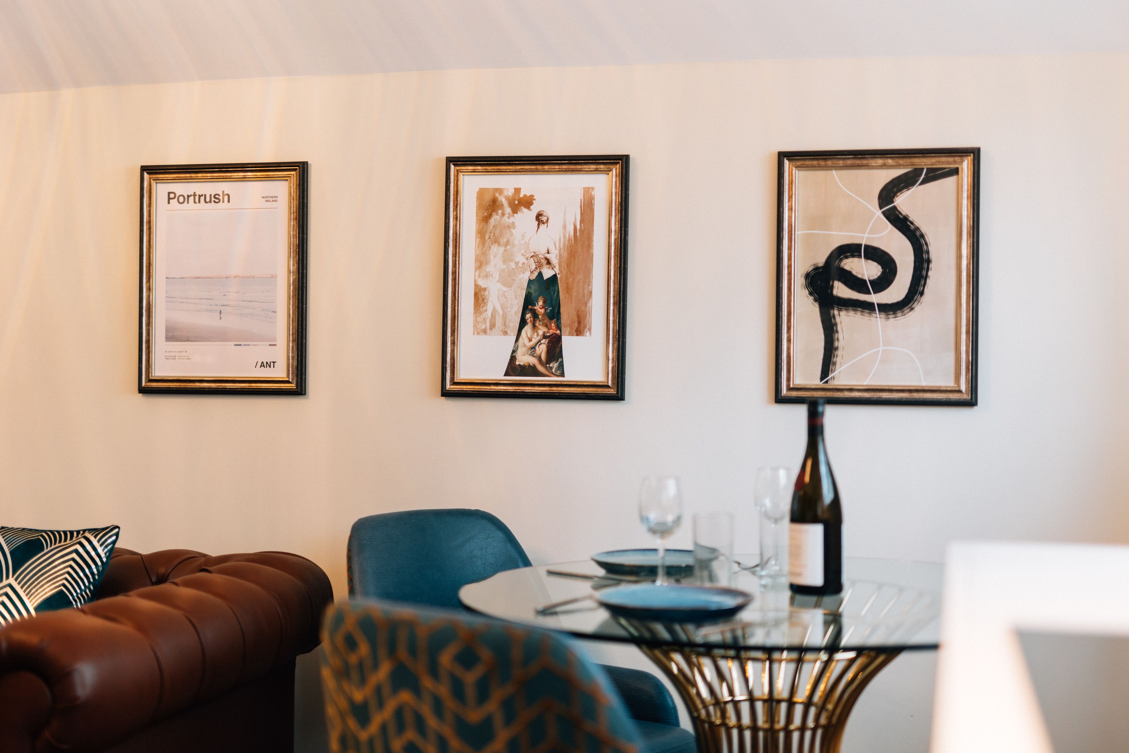 A dining area with a glass table, blue chairs, a brown leather couch, and framed artwork on the wall.