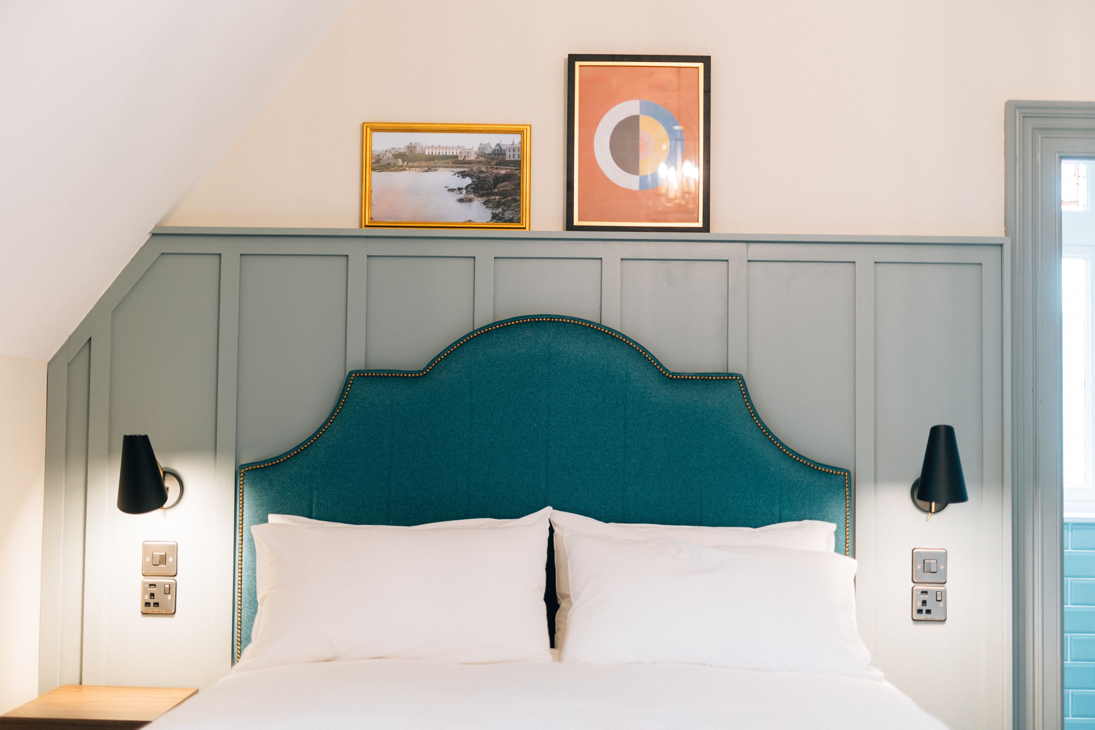 A neatly made bed with a teal headboard, two wall-mounted lamps, and two framed pictures on the wall above.