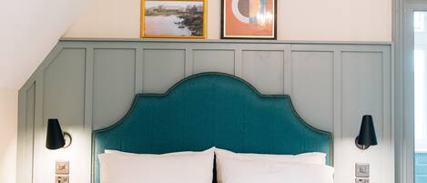 A neatly made bed with a teal headboard, two wall-mounted lamps, and two framed pictures on the wall above.