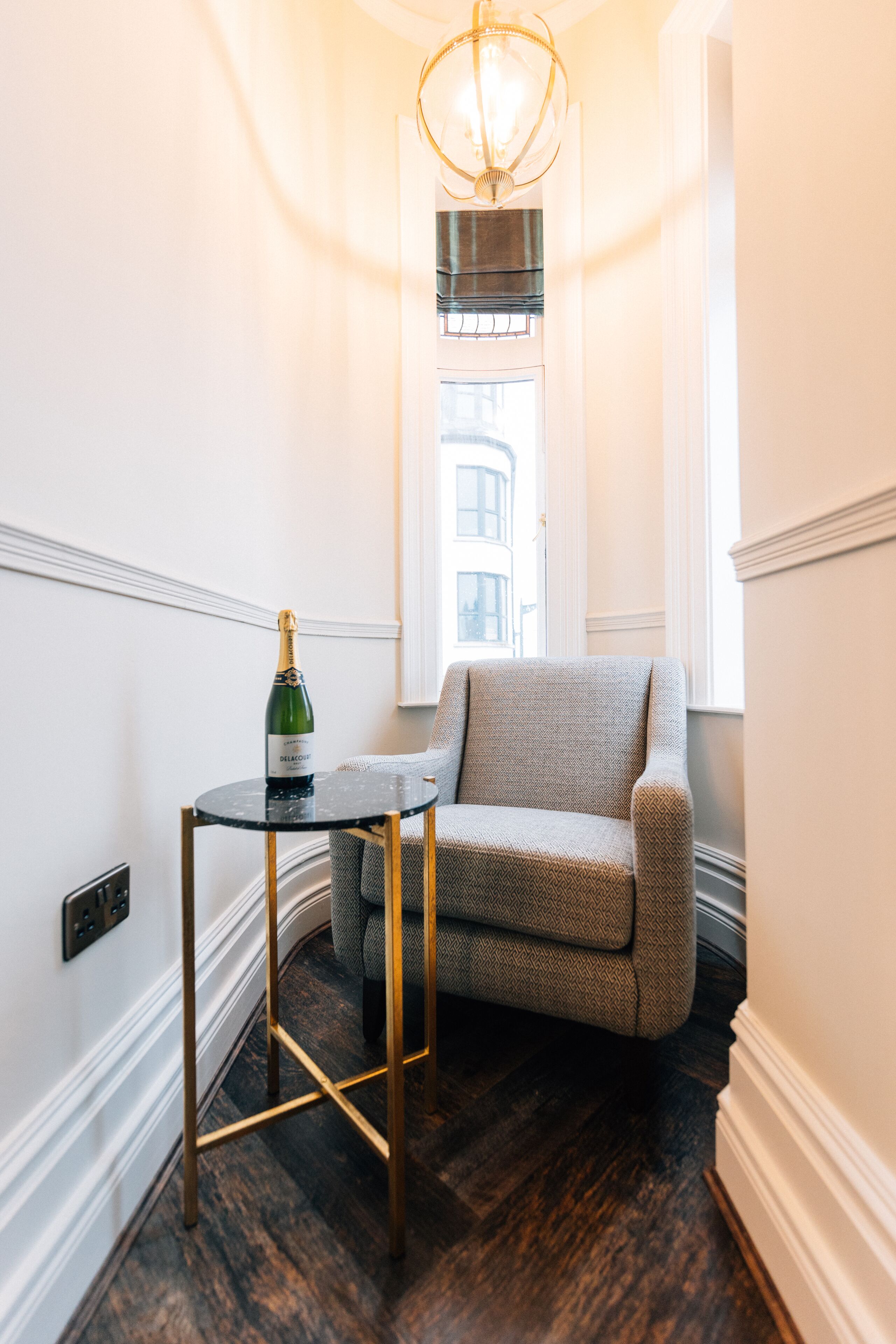 Une pièce d’angle avec un parquet en bois foncé, un fauteuil gris, une petite table ronde sur laquelle se trouve une bouteille de champagne, et un lustre.