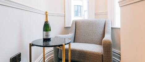 A corner room with a dark wood floor, a grey armchair, a small round table with a bottle of champagne, and a chandelier.