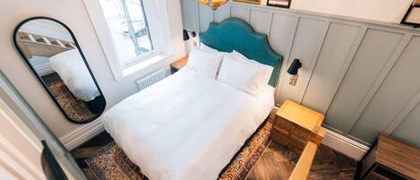 A neatly made bed with a headboard, a bedside table, a nightstand, a mirror, and a patterned rug.