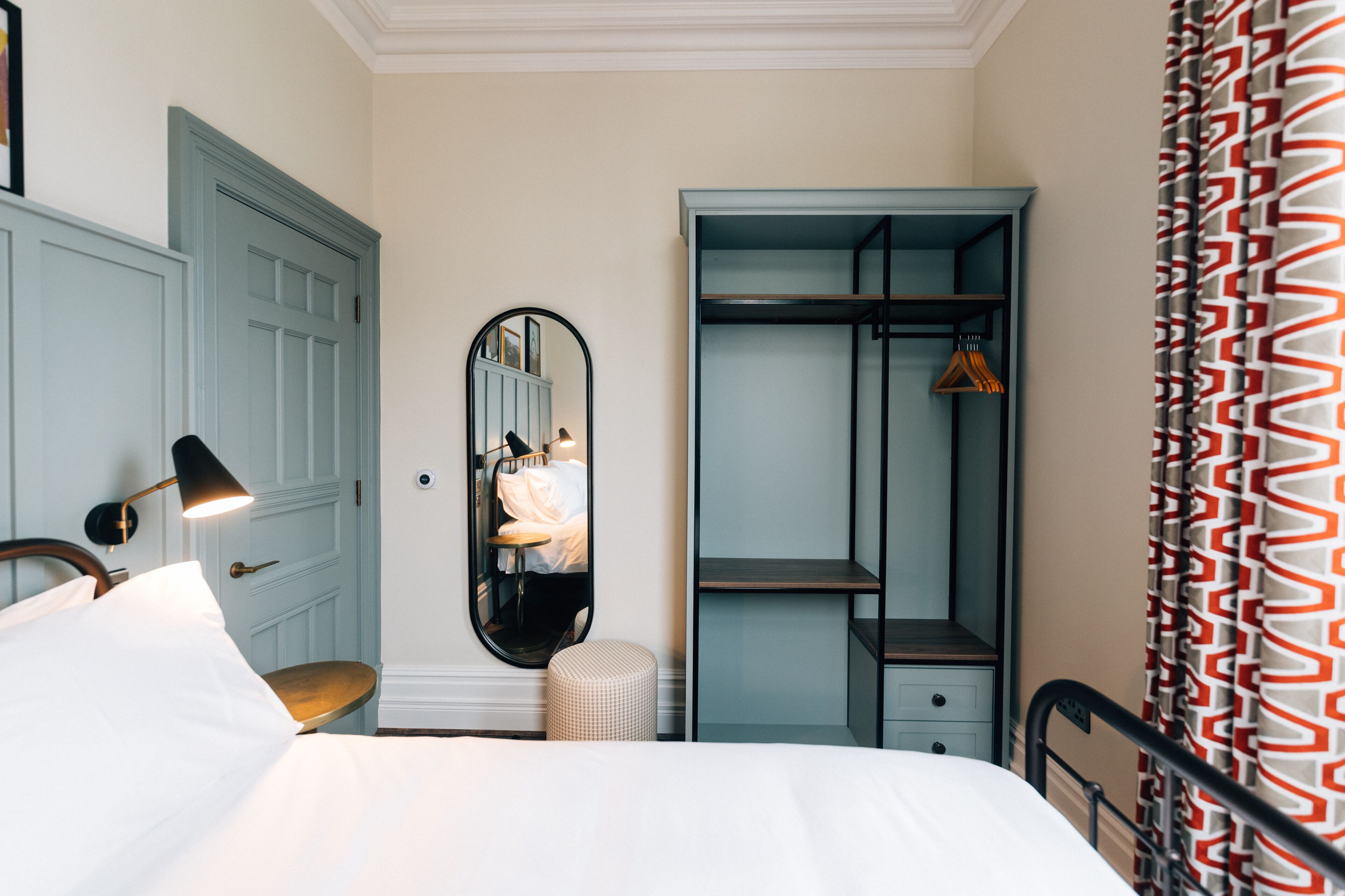 A hotel room with a bed, a bedside lamp, a mirror, a wardrobe, and a hanging coat rack.