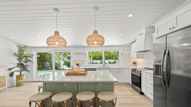 Cocina moderna con armarios blancos, una isla grande con taburetes de bar, electrodomésticos de acero inoxidable y amplias ventanas que ofrecen vistas a la vegetación.