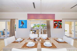 A dining area with a large table set for six, surrounded by white chairs, with artwork on the walls and a view of the outdoors.