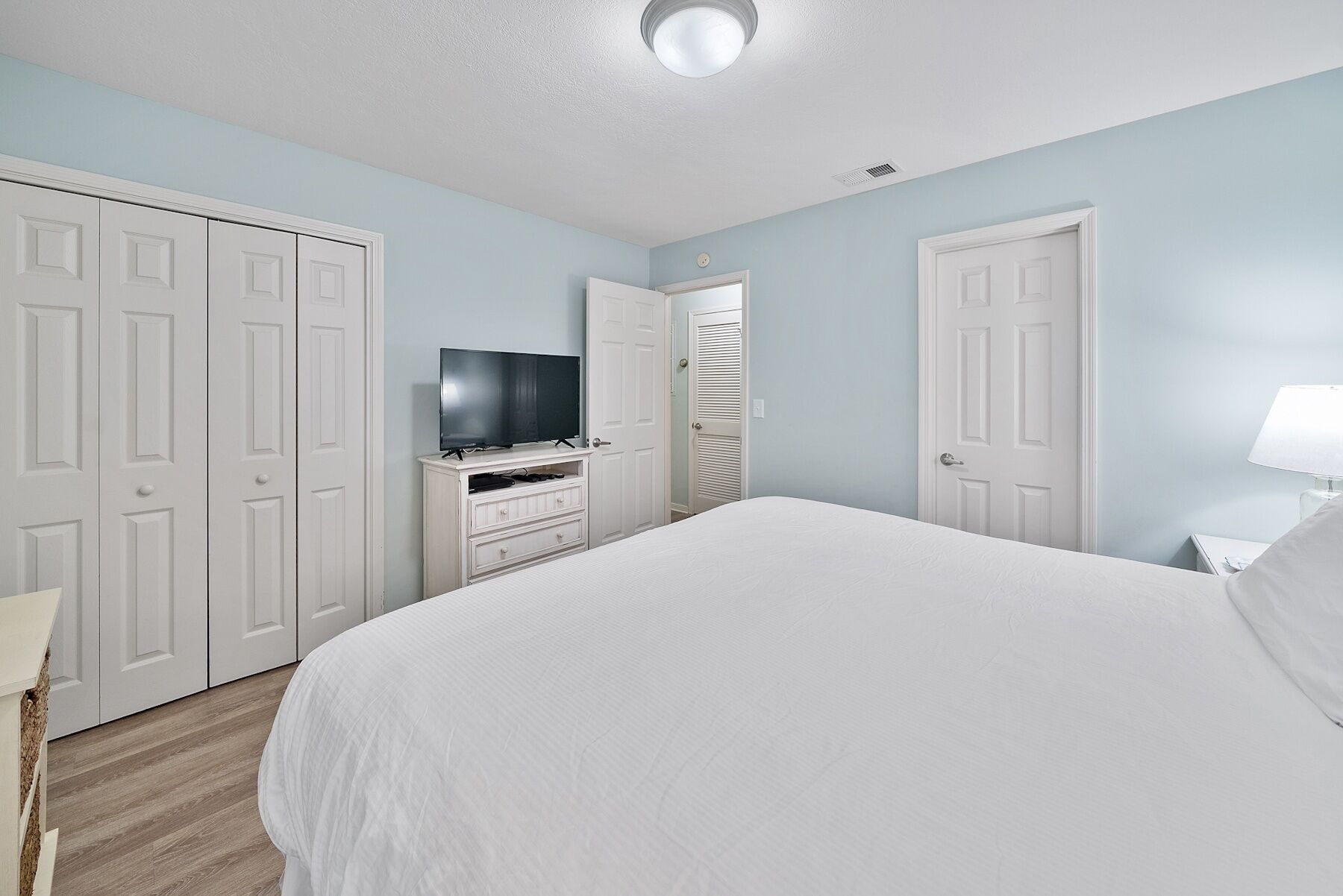 Ein Schlafzimmer mit einem großen Bett, einem weißen Kleiderschrank, einem Fernseher auf einem Standfuß und einer Tür mit Lamellenfenster.