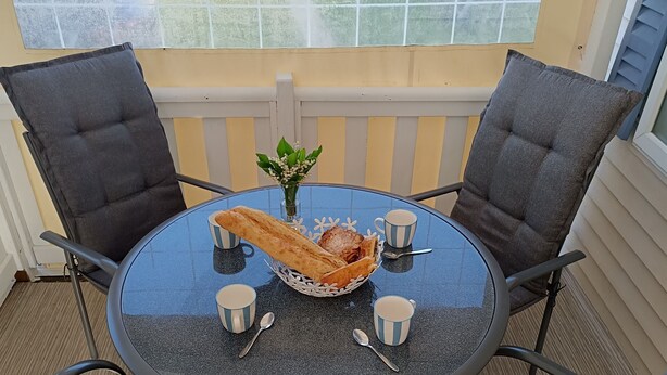 Ein runder Tisch mit einem Brotkorb, zwei Tassen und zwei Löffeln auf einem Balkon mit Stühlen und einem Geländer.