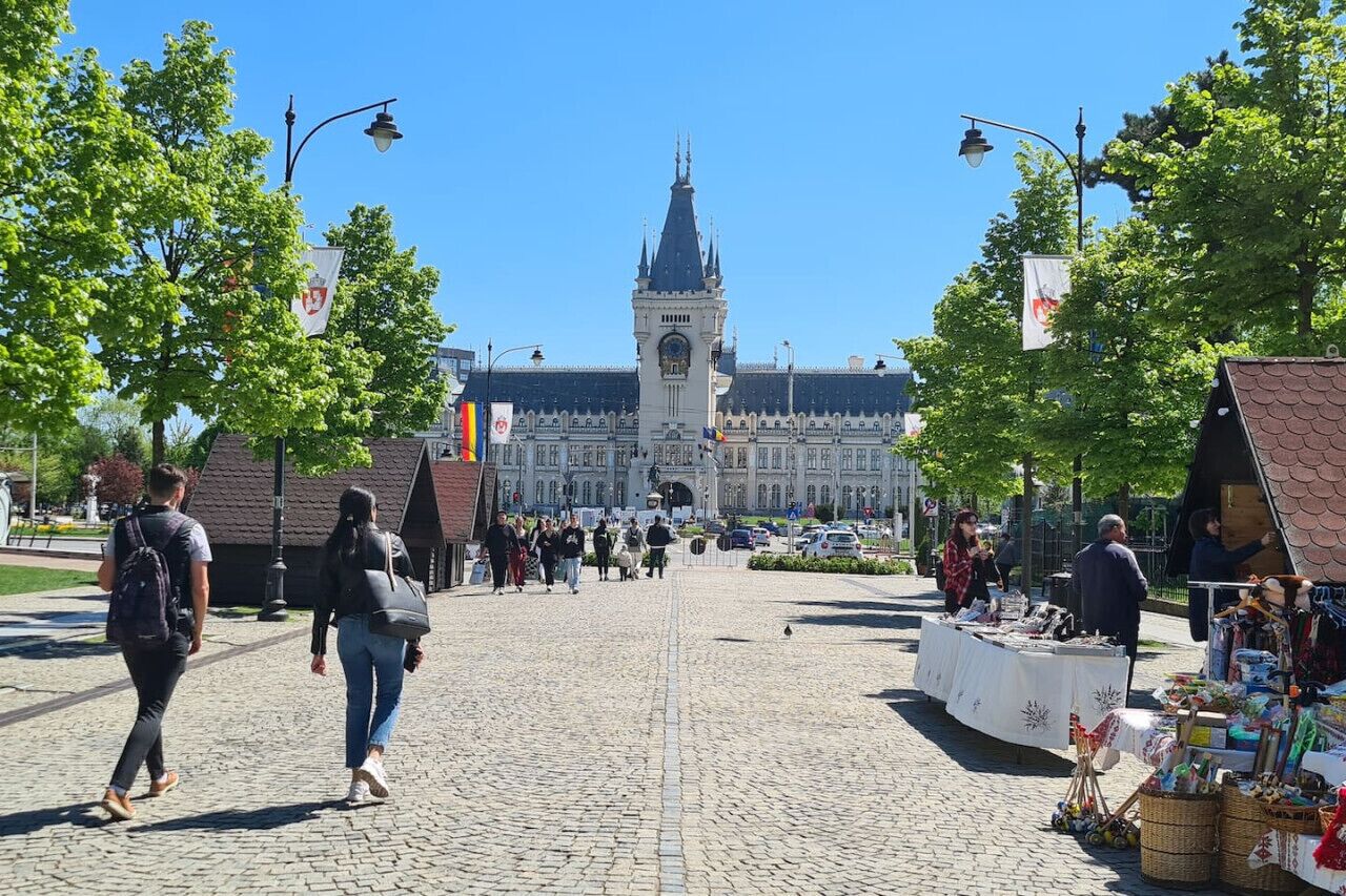 A cobblestone street with a large building in the background, a market stall on the right, and people walking.