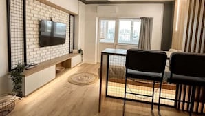 Un salon moderne avec un téléviseur à écran plat fixé à un mur en briques, un coin repas avec une table et des chaises, et une grande fenêtre avec des rideaux.