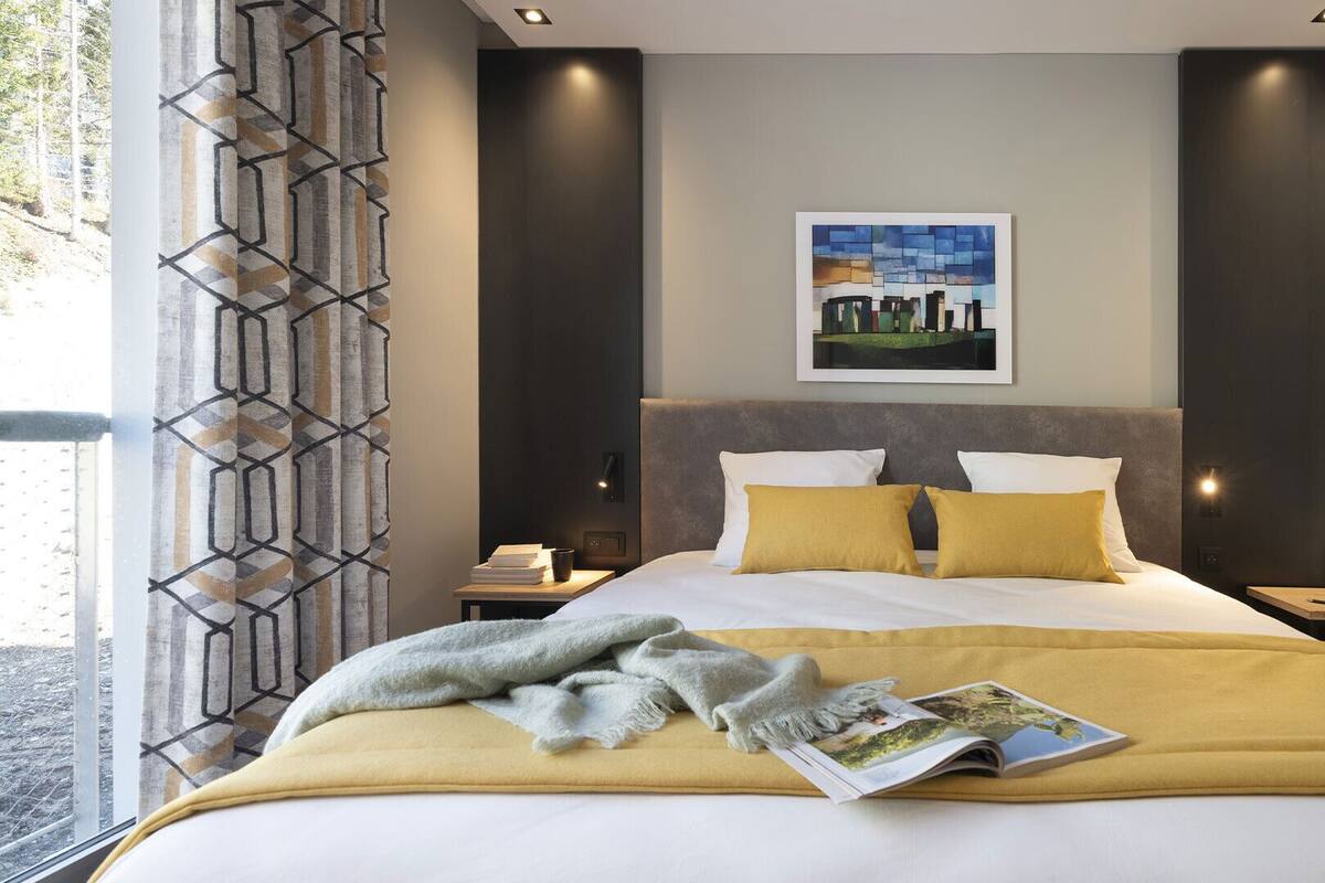 Una habitación de hotel moderna con una cama grande, dos mesitas de noche, una lámpara de noche, una obra de arte enmarcada sobre la cama y una ventana con cortina geométrica.